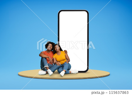 Sweet cheerful loving young black couple in casual sitting on round platform by huge phone with white blank screen mockup over blue background. Entertaining mobile app, blank space 115637863