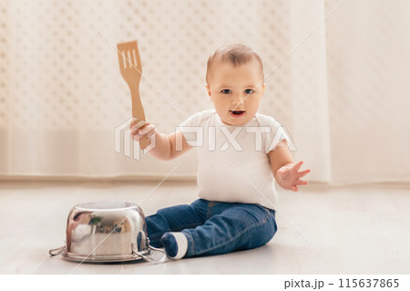 A small baby is seated on the floor, gripping a spatula in their tiny hands. The infant seems curious and engaged as they explore the kitchen utensil. A small baby is seated on the floor, gripping a spatula in their tiny hands. The infant seems curious and engaged as they explore the kitchen utensil. 115637865