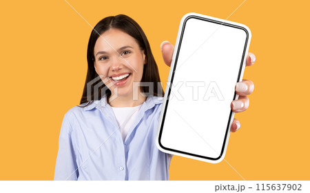 A young woman with long dark hair smiles broadly as she holds a smartphone with a blank white screen towards the camera. She is standing against a bright yellow background. 115637902