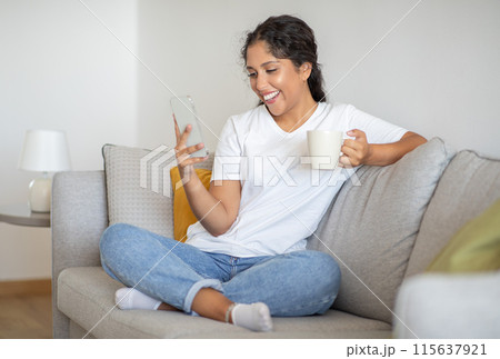 A woman sits on a light-colored sofa in a living room. She wears a white t-shirt and blue jeans and smiles while looking at her phone with a cup of coffee in her hand. 115637921