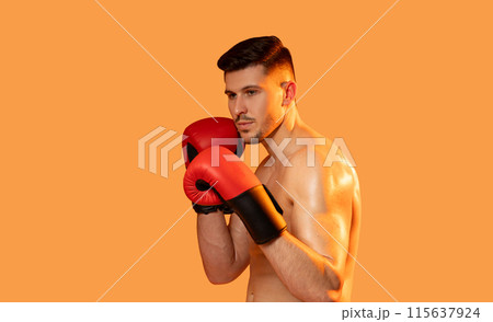 A man wearing boxing gloves strikes a pose against a vibrant orange background. He exudes confidence and strength as he showcases his boxing stance. A man wearing boxing gloves strikes a pose against a vibrant orange background. He exudes confidence and strength as he showcases his boxing stance. 115637924