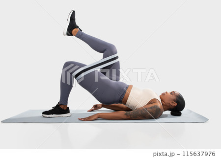 African American woman in athletic wear is doing a glute bridge exercise with a resistance band on a yoga mat. She is lying on her back with her knees bent and her feet flat on the mat. 115637976