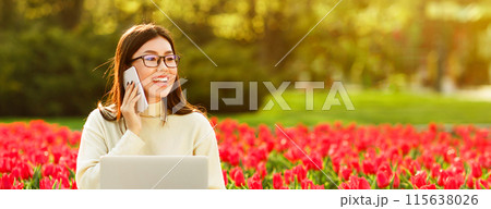 A cheerful Chinese woman chats on her phone while sitting among a field of bright red tulips. The sunlight enhances the lively atmosphere of the spring day. A cheerful Chinese woman chats on her phone while sitting among a field of bright red tulips. The sunlight enhances the lively atmosphere of the spring day. 115638026