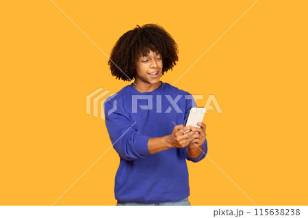 A young man with curly hair is wearing a blue shirt and smiling as he holds a phone in his hands. The backdrop is a vivid orange color which enhances the cheerful atmosphere of the scene. A young man with curly hair is wearing a blue shirt and smiling as he holds a phone in his hands. The backdrop is a vivid orange color which enhances the cheerful atmosphere of the scene. 115638238