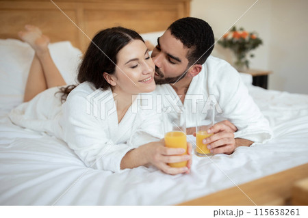 Husband Kissing While Wife They Sip Orange Juice, Having Fresh Morning Drink And Romantic Breakfast In Bed At Luxury Hotel. Spouses Relaxing Enjoying Great Room Service At Vacation Accomodation 115638261