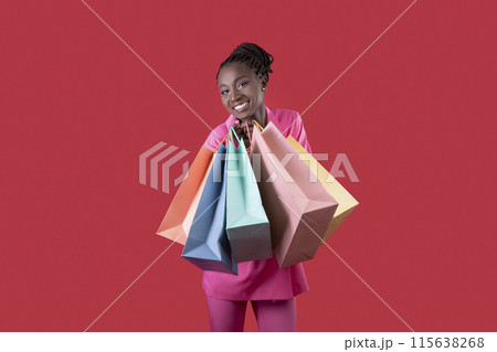 Beautiful Smiling African American Woman With Shopping Bags In Hands Posing Over Red Studio Background, Happy Black Shopaholic Lady In Pink Suit Looking At Camera, Enjoying Seasonal Sales, Copy Space 115638268