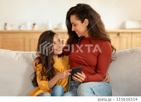 Happy Middle Eastern Mom And Little Daughter Using Smartphone, Kid Girl Pointing Finger At Gadget, Showing Her Favorite Mobile Game Application, Hugging Sitting On Couch At Home Interior Happy Middle Eastern Mom And Little Daughter Using Smartphone, Kid Girl Pointing Finger At Gadget, Showing Her Favorite Mobile Game Application, Hugging Sitting On Couch At Home Interior 115638314