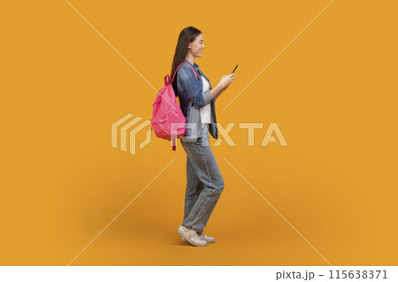 A young woman with long dark hair is wearing a blue jean jacket and jeans. She is carrying a pink backpack and using a smartphone. She is standing on a yellow background. 115638371