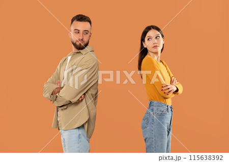 A young man and woman are standing back to back, both with arms crossed, indicating disagreement or conflict. The background is a solid orange color A young man and woman are standing back to back, both with arms crossed, indicating disagreement or conflict. The background is a solid orange color 115638392