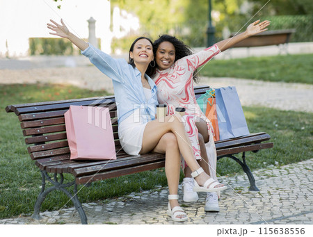 Two Beautiful Young Woman With Shopping Bags Posing On Bench Outdoors, Happy Cheerful Multiethnic Female Friends Having Fun Together, Drinking Takeaway Coffee, Enjoying Seasonal Sales 115638556