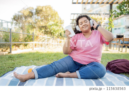 Happy Overweight Latin Woman Listening To Music Online With Headphones And Smartphone, Sitting On Blanket Enjoying Picnic In Park Outside. Lady Closing Eyes, Listens Favorite Playlist On Summer Day 115638563