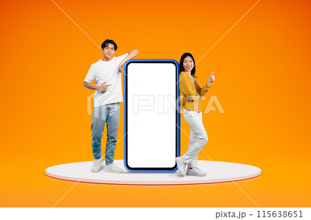 Cheerful chinese millennials casual man and woman posing on platform by big phone with white empty screen, showing thumb up, isolated on orange background, blank space. Great online offer 115638651