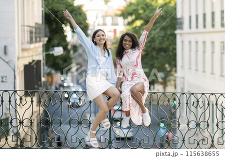 Two Cheerful Young Female Friends Having Fun Together Outdoors, Happy Multiethnic Besties Laughing And Gesturing While Sitting On City Fence, Raising Hands And Looking At Camera, Free Space Two Cheerful Young Female Friends Having Fun Together Outdoors, Happy Multiethnic Besties Laughing And Gesturing While Sitting On City Fence, Raising Hands And Looking At Camera, Free Space 115638655