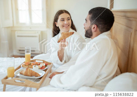 Loving Mornings. Joyful Wife Offering Croissant To Husband During Breakfast In Bed, Enjoying Each Other's Company With Tray Of Food In Hotel Bedroom Interior. Honeymoon Vacation Pleasures 115638822