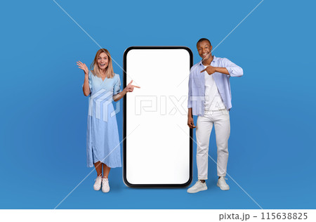 A man and woman are pointing at a blank screen against a solid blue background. Both individuals are gesturing towards the screen, displaying engagement and interaction. 115638825