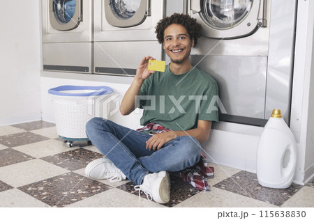 Young Latin Man Showing Credit Card Sitting With Detergent Bottle At Laundry Room Indoor, Smiling At Camera Posing Near Row Of Industrial Washing Machines. Great Public Conveniences Offer Young Latin Man Showing Credit Card Sitting With Detergent Bottle At Laundry Room Indoor, Smiling At Camera Posing Near Row Of Industrial Washing Machines. Great Public Conveniences Offer 115638830