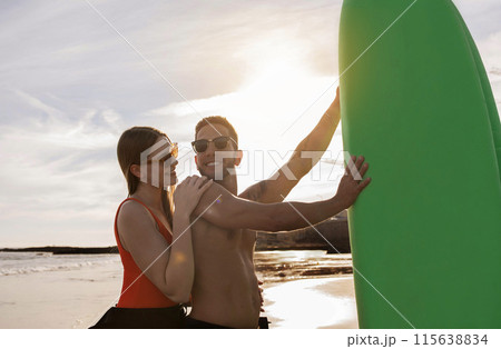 Sporty Surfers Couple Having Fun While Surfing Together On The Beach At Sunset, Happy Millennial Man And Woman Posing With Surfboard, Wearing Stylish Eyeglasses And Wetsuits, Enjoying Water Sports Sporty Surfers Couple Having Fun While Surfing Together On The Beach At Sunset, Happy Millennial Man And Woman Posing With Surfboard, Wearing Stylish Eyeglasses And Wetsuits, Enjoying Water Sports 115638834