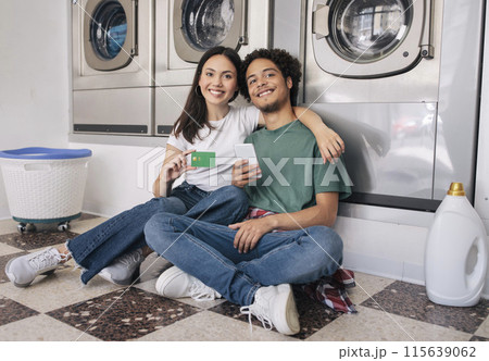 Laundry Offer. Happy Young Couple Showing Credit Card Sitting Near New Washing Machines, Using Smartphone Enjoying Self Service And Smiling To Camera. Enjoy Household Leisure Together 115639062