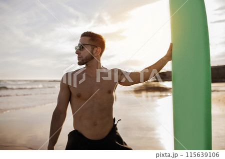 Portrait of handsome young man holding surfboard at the beach, professional surfer guy in stylish eyeglasses posing on sunset, shirtless millennial male standing on ocean shore, ready for surfing 115639106