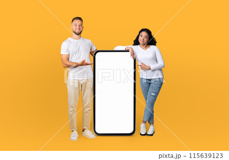 A man and a woman are standing together, holding a blank phone in their hands. They are posing against a bright yellow background, looking directly at the camera. A man and a woman are standing together, holding a blank phone in their hands. They are posing against a bright yellow background, looking directly at the camera. 115639123