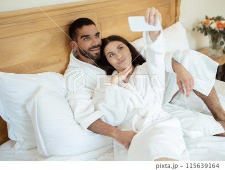 Happy loving couple resting in hotel room and taking selfie via mobile phone, lying in bed and posing wearing bathrobes, having fun on honeymoon vacation. Gadgets and social media concept Happy loving couple resting in hotel room and taking selfie via mobile phone, lying in bed and posing wearing bathrobes, having fun on honeymoon vacation. Gadgets and social media concept 115639164