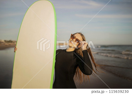 Portrait Of Beautiful Young Surfer Lady Holding Big Blank Surfboard, Smiling Millennial Woman Wearing Stylish Sunglasses And Wetsuit Standing On The Beach At Sunset, Enjoying Surfing, Copy Space Portrait Of Beautiful Young Surfer Lady Holding Big Blank Surfboard, Smiling Millennial Woman Wearing Stylish Sunglasses And Wetsuit Standing On The Beach At Sunset, Enjoying Surfing, Copy Space 115639230