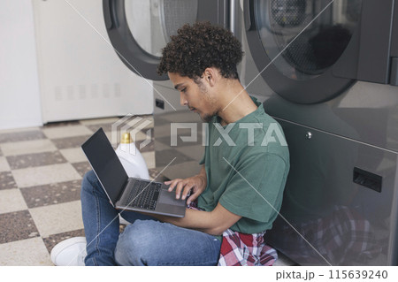 Great Laundry Service. Middle Eastern Student Guy Learning Online Via Laptop While Washing Clothes At Public Laundrette, Sitting Indoor With Computer And Surfing Web, Wearing Casual. Side View 115639240