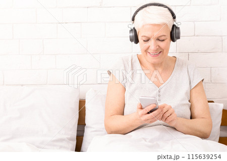 Senior woman listening to music on his smartphone at home in bed, using modern wireless headphones, panorama, copy space Senior woman listening to music on his smartphone at home in bed, using modern wireless headphones, panorama, copy space 115639254