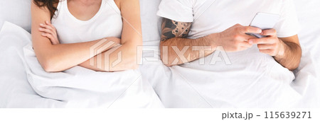A woman with her arms crossed lies in a bed beside a man who is using his phone. She appears upset or disinterested, while the man is engrossed in the screen, cropped, panorama A woman with her arms crossed lies in a bed beside a man who is using his phone. She appears upset or disinterested, while the man is engrossed in the screen, cropped, panorama 115639271