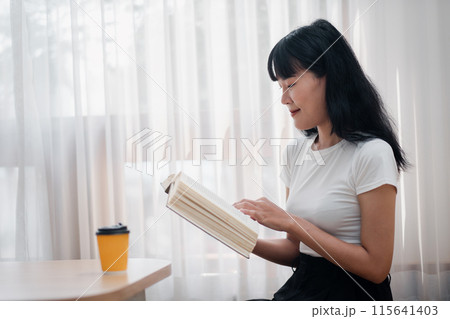 A young woman in a white shirt reads a book by the window with a coffee cup on the table, enjoying a peaceful moment. 115641403