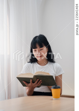 A young woman in casual clothing reading a book at home with a cup of coffee on a wooden table, enjoying a peaceful and relaxed atmosphere. A young woman in casual clothing reading a book at home with a cup of coffee on a wooden table, enjoying a peaceful and relaxed atmosphere. 115641404