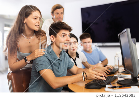 Girl stands behind sitting students, looks at monitor and helps in execution of programing teamwork Girl stands behind sitting students, looks at monitor and helps in execution of programing teamwork 115642083