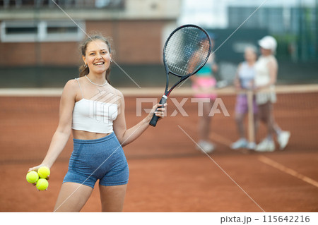 Cheerful girl with racket and tennis balls standing on outdoor court 115642216