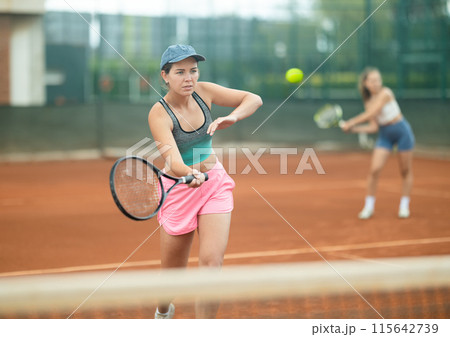 Girl swinging racket to hit ball during tennis doubles on outdoor court 115642739