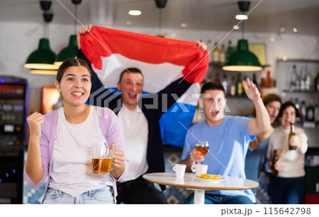 Company of excited young adult sports fans waving Dutch flag and supporting national team with beer in pub 115642798
