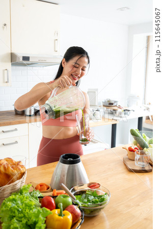 An attractive, healthy Asian woman in sportswear is pouring her green juice from a blender. 115645067