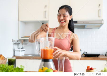 A fit Asian woman in sportswear making a smoothie or healthy juice in the kitchen with a blender. A fit Asian woman in sportswear making a smoothie or healthy juice in the kitchen with a blender. 115645090