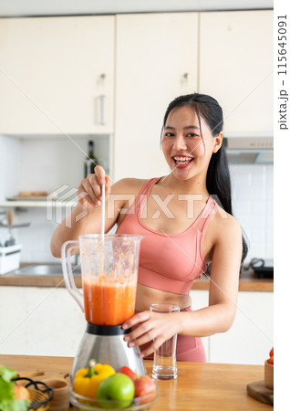 A fit Asian woman in sportswear making a smoothie or healthy juice in the kitchen with a blender. 115645091
