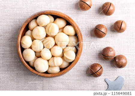 Roasted macadamia nuts in a wooden bowl on linen fabric. On the right fruits with sawn nutshells and opener key. Also known as Queensland, bush, maroochi, bauple and Hawaii nuts. Close-up, from above. 115649081