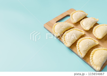 Raw samosas with sesame on a wooden on board on blue table ready for baking. Traditional Central Asian cuisine, frozen semi-finished products, copy space. Raw samosas with sesame on a wooden on board on blue table ready for baking. Traditional Central Asian cuisine, frozen semi-finished products, copy space. 115654867