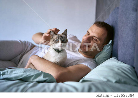 a  young man pet a cat in bed, morning routine 115655384