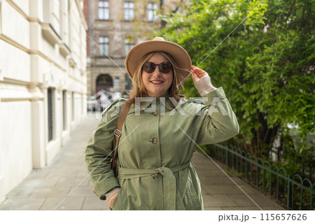 Portrait of a beautiful Caucasian female in a green coat, carrying a walking bag, smiling in a good mood in the city outdoors. Portrait of a beautiful Caucasian female in a green coat, carrying a walking bag, smiling in a good mood in the city outdoors. 115657626