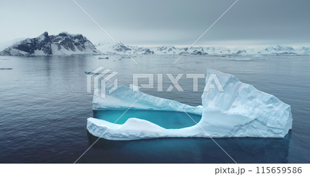 Antarctic melting iceberg floating in cold blue water, aerial. Snow-covered mountains in background. Melting glacier drift in Polar ocean. Ecology, melting ice, climate change, global warming concept Antarctic melting iceberg floating in cold blue water, aerial. Snow-covered mountains in background. Melting glacier drift in Polar ocean. Ecology, melting ice, climate change, global warming concept 115659586