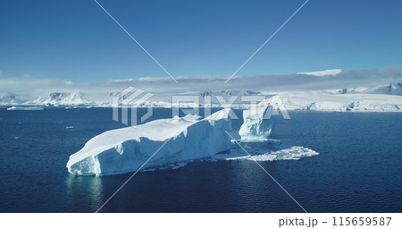 Huge blue iceberg drifts arctic ocean, clear sunny sky. Towering snow covered glacier melting in polar water. Mountains in background. Global warming issue of climate change and melting icebergs 115659587