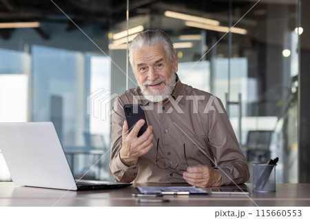 Senior businessman working on laptop and mobile phone in modern office while looking at camera and smiling Senior businessman working on laptop and mobile phone in modern office while looking at camera and smiling 115660553