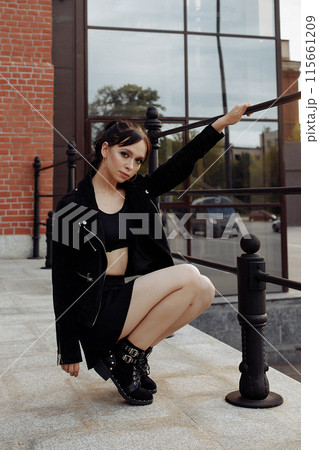 A young woman in black poses for a photo. She is wearing black boots, a jacket and a miniskirt. A young woman in black poses for a photo. She is wearing black boots, a jacket and a miniskirt. 115661209