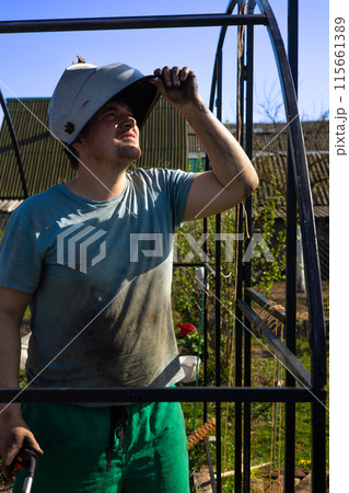 A dirty man in a protective mask inspects the welding site. The welder takes off his mask to check the result of his work. In the garden, a man welds the metal frame of a greenhouse. A dirty man in a protective mask inspects the welding site. The welder takes off his mask to check the result of his work. In the garden, a man welds the metal frame of a greenhouse. 115661389
