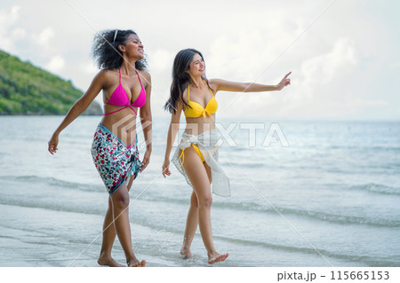 Two women are walking on the beach, one wearing a pink bikini Two women are walking on the beach, one wearing a pink bikini 115665153