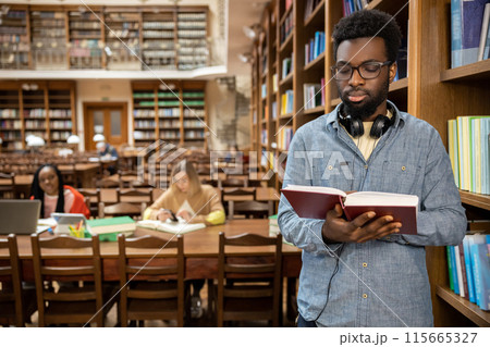 Dark-skinned young man in eyeglasses with a book in hands in the library Dark-skinned young man in eyeglasses with a book in hands in the library 115665327