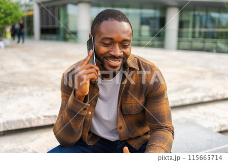 African american man talking on smartphone on street in city outdoor. Man with cell phone chatting with friends. Smiling person making answering call by cellphone. Guy having conversation by mobile 115665714